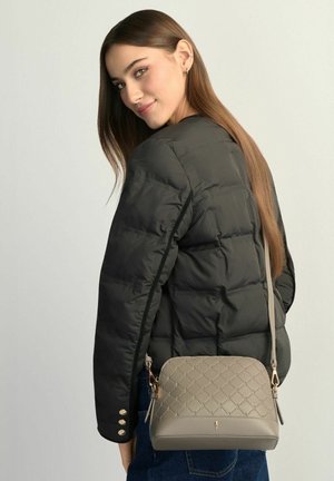 Młoda kobieta o długich brązowych włosach, ubrana w czarną pikowaną kurtkę i niosąca beżową pikowaną torebkę na ramię.