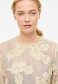 Haut beige clair transparent avec broderie florale en motif texturé, encolure ronde ornée d'une délicate bordure en dentelle, et manches longues et fluides.