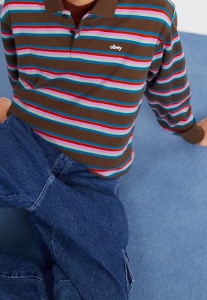 Braun gestreiftes Polohemd mit pinken, blauen und roten horizontalen Linien, kombiniert mit blauen Denim-Jeans. Das Hemd trägt ein "obey"-Logo auf der Brust.