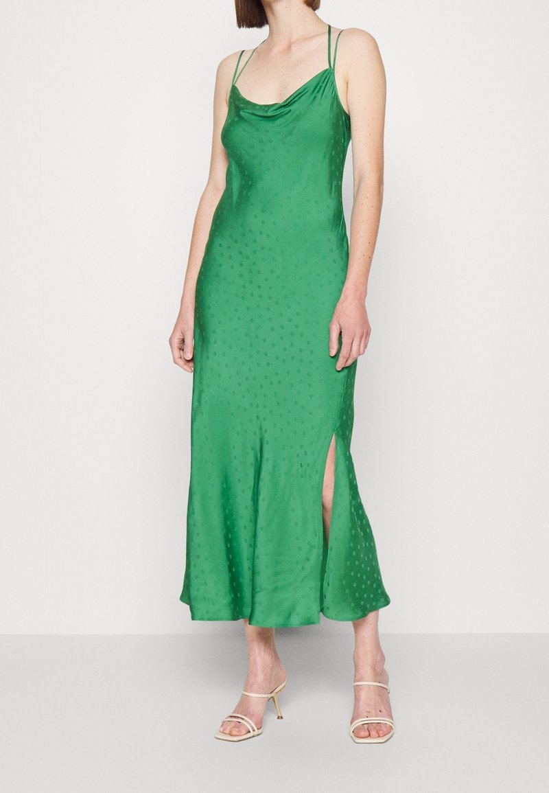 Femme portant une robe nuisette midi verte sans manches à pois avec une fente latérale, associée à des sandales à talons hauts beiges.