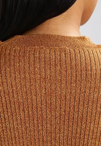 Sweter z prążkowanego dzianiny w ciepłych, złoto-brązowych odcieniach. Posiada okrągły dekolt z teksturowanym wykończeniem i delikatnym połyskiem, podkreślającym detal tkaniny.