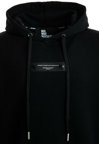 Sudadera negra con capucha que presenta una tela texturizada, cordones con puntas plateadas y una etiqueta rectangular negra con texto blanco en el frente.