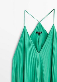 Robe plissée verte sans manches avec fines bretelles et un décolleté en V profond, suspendue sur un fond blanc.