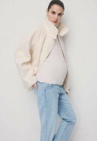 Kobieta w ciąży ubrana w kremowy płaszcz teddy, jasny sweter i niebieskie dżinsy, stojąca z jedną ręką w kieszeni na jednolitym tle.