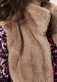 Kobieta nosząca jasnobrązowy, pluszowy szal z futra syntetycznego na wzorzystej bluzce w czarnym, różowym i białym kolorze oraz z luźnymi, falującymi brązowymi włosami.