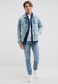Chaqueta de denim azul claro con dos bolsillos en el pecho, superpuesta a una camisa azul oscura y una camiseta blanca. Combinada con jeans azul claro y zapatillas blancas.