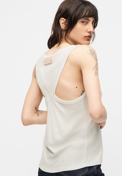 Άτομο με κοντά σκούρα μαλλιά που φοράει άσπρη αμάνικη μπλούζα, δείχνοντας την πλάτη και τα τατουάζ στο χέρι και τον ώμο, κοιτάζοντας πάνω από τον ώμο.