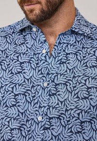 Camisa de manga corta en azul claro y marino con un patrón de hojas, cuello abotonado y textura suave. Cinco botones visibles en la tapeta.