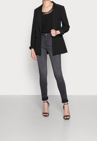Blazer negro sobre una blusa negra con un escote brillante, combinado con jeans skinny gris oscuro y tacones negros con tiras.