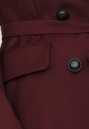 Cappotto in tessuto bordeaux con colletto a punta, grandi bottoni neri e cintura in vita. Presenta una tasca laterale e una texture liscia.