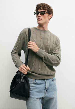 Mann trägt einen grauen Zopfpullover, hellblaue Jeans, schwarze Sonnenbrille und hält eine schwarze Lederschultertasche.