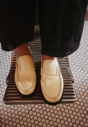 Pieds portant des mocassins couleur crème avec des détails cousus, debout sur une marche métallique à lattes au-dessus d'un sol carrelé hexagonal.