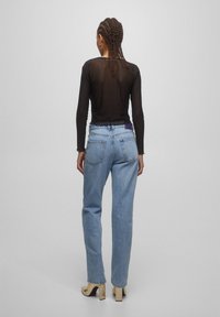 Haut noir transparent à manches longues associé à un jean bleu clair taille haute et des chaussures à talons beiges, vu de dos.