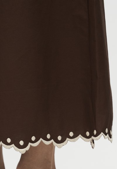 Καφέ πατούρα φούστας με λευκή κεντητή διακόσμηση σε κυματιστό σχέδιο πάνω από γυμνά πόδια σε ανοιχτό φόντο.