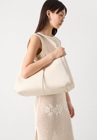 Cream-colored hobo bag made from structured fabric, featuring subtle gold-tone hardware and a spacious design, held by a model.