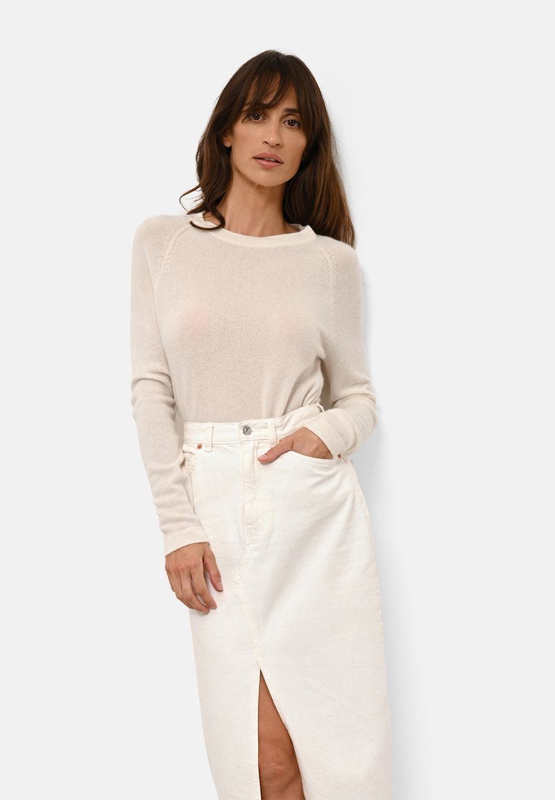 Pull en maille beige clair avec des manches longues, un col rond et des poignets côtelés, associé à une jupe blanche taille haute avec une fente à l'avant.