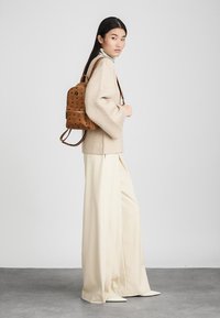 Kvinna klädd i beige vida byxor, en oversized beige tröja och en brun mönstrad läderryggsäck, stående på sidan mot en enfärgad bakgrund.