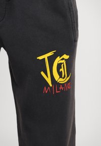 Svarte joggebukser med en myk tekstur, med et gult og rødt grafisk design som leser "JC Milano" i en fet, stilisert skrift.