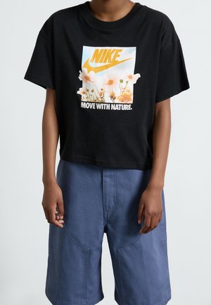 Person iført en sort Nike T-shirt med orange logo og blomster, tekst "MOVE WITH NATURE," kombineret med blå shorts.