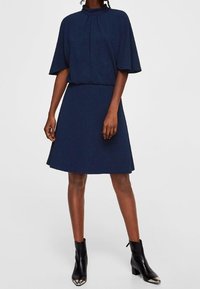 Robe bleu marine à manches courtes avec un col haut et un détail de pli, présentant une taille ajustée et une jupe évasée. Portée avec des bottines.