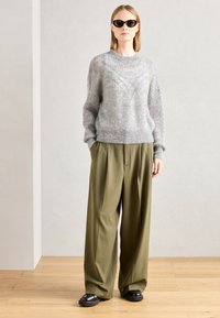 Grauer Strickpullover mit gerippter Textur und V-Muster, kombiniert mit lockeren olivgrünen Hosen und schwarzen klobigen Schuhen. Das Modell trägt Sonnenbrillen.
