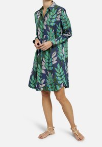 Abito a camicia con un motivo verde foglia e blu navy. Maniche lunghe, vestibilità rilassata, lunghezza sopra il ginocchio. Indossato con sandali con cinturino di colore chiaro.