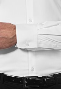 Chemise blanche à manches longues avec col classique, poignets structurés et fermeture à boutons. Ceinture en cuir noir avec une boucle rectangulaire brillante.