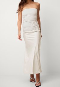 Axelbandslös klänning i krämfärg med rynkad överdel och mjuk, golvlång kjol; kombinerad med silverfärgade flip-flops och minimala detaljer.