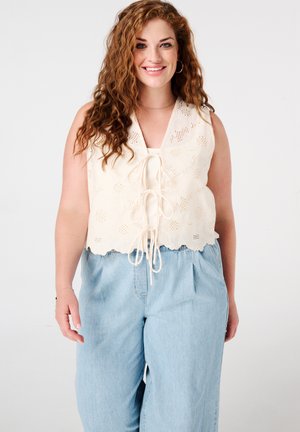 Witte bloemenkant crop top met een V-hals en striksluiting aan de voorkant, gecombineerd met lichtblauwe high-waisted wijde jeans.
