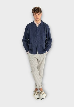 Veste en coton bleu marine avec deux poches avant, fermeture à boutons et coupe décontractée, associée à un pantalon à rayures et des baskets blanches.