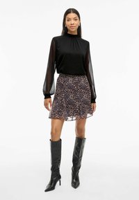 Blusa nera a maniche lunghe con scollatura volant, abbinata a una gonna corta e fluida con un motivo floreale scuro. Stivali neri fino al ginocchio.