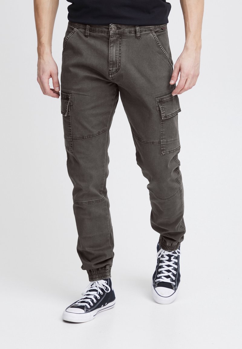 Homme portant un pantalon cargo gris foncé avec des poignets élastiques et des baskets montantes noires à lacets blancs sur un fond blanc.