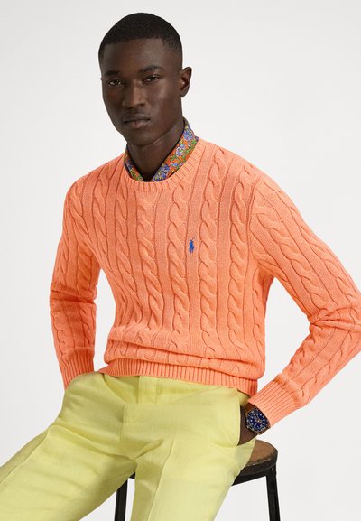 Cable-knit orange sweater with long sleeves, crew neck, and blue logo. Worn over a patterned collar shirt, paired with light yellow pants.