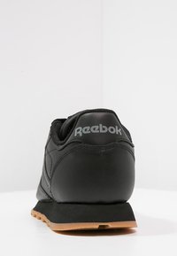 Svart lær-sneaker med teksturert overflate og tan gummisåle. Har Reebok-logoen trykket i grått på hælen.