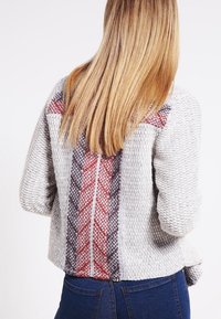 Šedý pletený svetr s vzorovanými zády v červené a tmavě šedé barvě, s kulatým lemem a spadlými rameny.