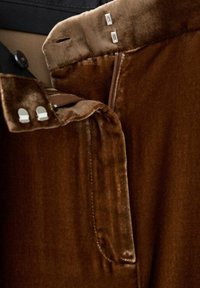 Giacca in velluto marrone con una texture liscia, caratterizzata da una zip, dettagli metallici e un design su misura. L'interno è di un tessuto più chiaro.