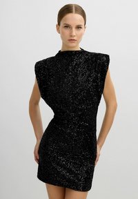 Vestido mini negro de lentejuelas con una silueta ajustada, cuello alto y hombros estructurados. Textura suave con adornos brillantes.