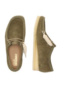 Gröna mockaskor med en mjuk textur, med en krämfärgad snörningsdetalj och en böjd gummisula. Enkel design med vita sömnadsdetaljer.