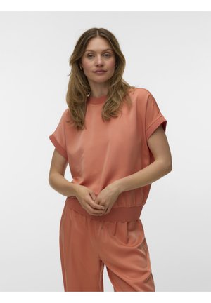 Vrouw met golvend lichtbruin haar draagt een losse koraalkleurige korte mouw top en bijpassende broek, poseren tegen een effen witte achtergrond.