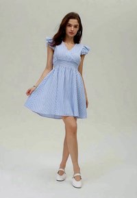 Abito corto a strisce blu e bianche con maniche a cappuccio, vita aderente e gonna svasata. Indossato con scarpe bianche e accessori minimi.
