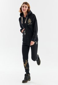 Čierna mikina na zips so zlatým logom Hogwarts a so šnúrkami, skombinovaná s čiernymi teplákmi s dizajnom zlatého jelena. Čierne boots dopĺňajú tento vzhľad.