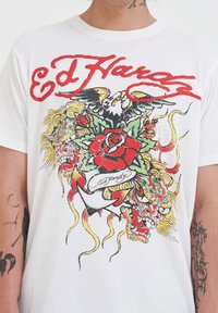 Camiseta blanca de algodón con un diseño colorido inspirado en tatuajes que incluye una rosa, un dragón y un águila, realzado por texto en rojo.