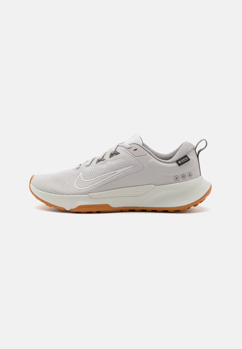 Zapatilla deportiva gris claro con parte superior de malla texturizada, suela de goma y acentos marrones. Presenta un logo y un lazo en el talón.
