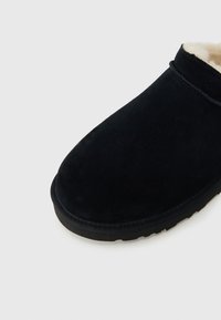 Μαύρο σουέτ παντόφλα με στρογγυλεμένο σχήμα, μαλακή εσωτερική επένδυση και υφασμένο καουτσούκ πάτο. Ελάχιστες ραφές τονίζουν την πάνω άκρη.