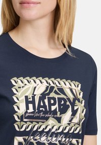 Granatowa koszulka z okrągłym dekoltem, ozdobiona nadrukiem liści oraz wyboldowanym napisem "SZCZĘŚLIWY" w jasnych i ciemnych kolorach.