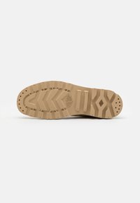 Semelle de botte beige avec des crampons profonds, motifs en chevrons en relief, perforations circulaires et nom de la marque embossé au centre.