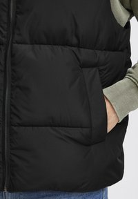 Schwarze Puffweste mit einem Reißverschluss, gerippten olivgrünen Bündchen und einer Fronttasche. Hat horizontale gesteppten Abschnitte für Isolierung.