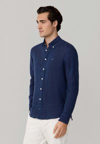 Man draagt een donkerblauw overhemd met witte knopen en witte broek, staand tegen een effen lichtgrijze achtergrond.