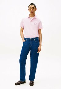 Roze poloshirt met kraag, blauwe denim jeans en bruine leren schoenen. Model staat met de hand in de zak voor een eenvoudige witte achtergrond.