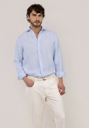 Hombre con cabello castaño lleva camisa de botones rayada azul claro y pantalones color crema con cinturón tejido, de pie con una mano en el bolsillo.